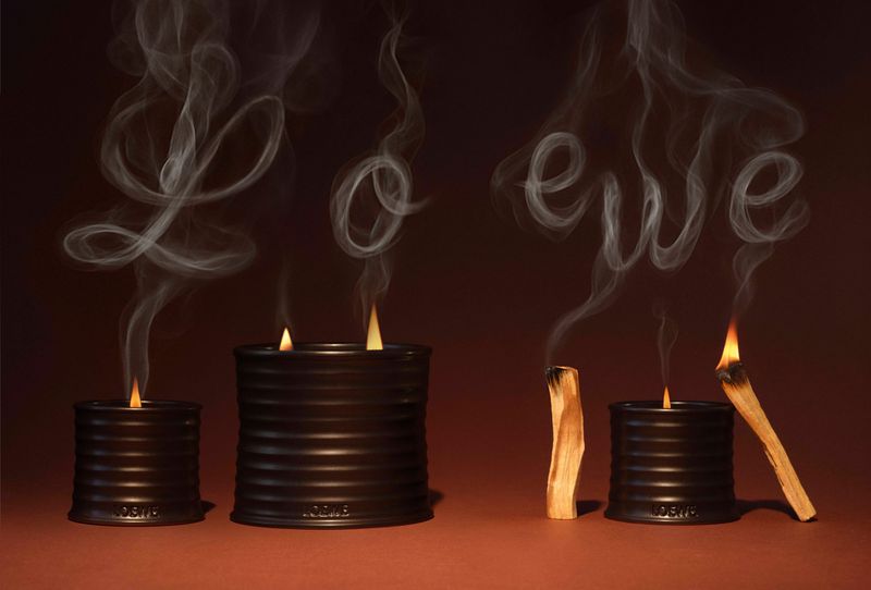 Three lit candles with smoke forming the word 'LOEWE' against a brown background create a moody, artistic ambiance in a still life image.