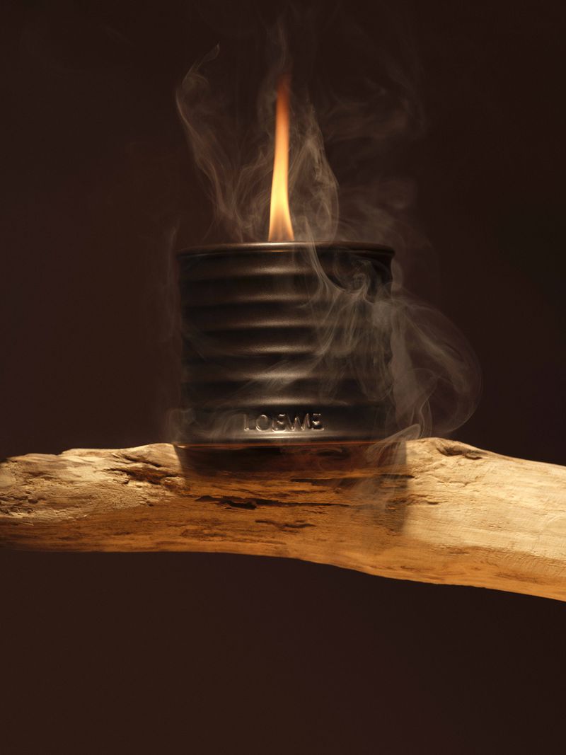 Candle flame burning in a Loewe candle holder sitting atop a piece of wood, surrounded by smoke, against a brown background.