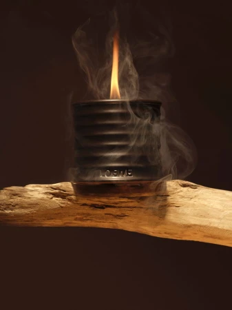 Candle flame burning in a Loewe candle holder sitting atop a piece of wood, surrounded by smoke, against a brown background.