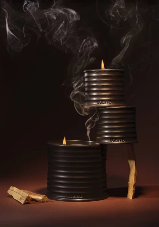 Three stacked dark ribbed candle tins with burning wicks and wisps of smoke against a warm brown background