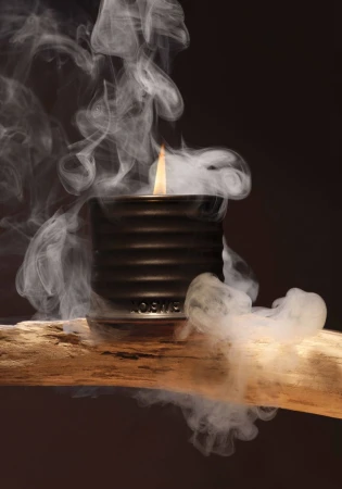 Lit black candle in a ribbed holder resting on a piece of rough wood with swirling smoke against a dark background.