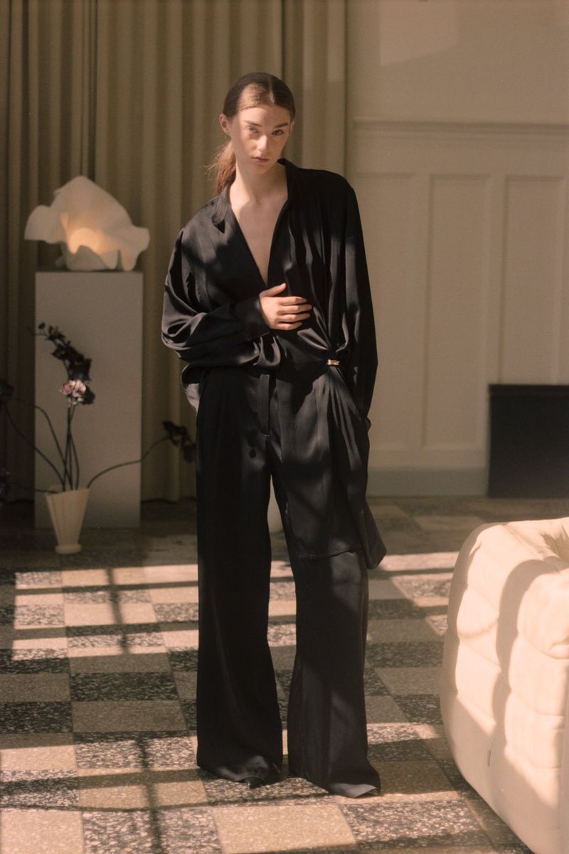 A slender woman in a draped black satin blouse and flowing black pants stands in a light-filled room with terrazzo tile floors.