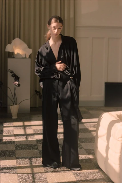 A slender woman in a draped black satin blouse and flowing black pants stands in a light-filled room with terrazzo tile floors.