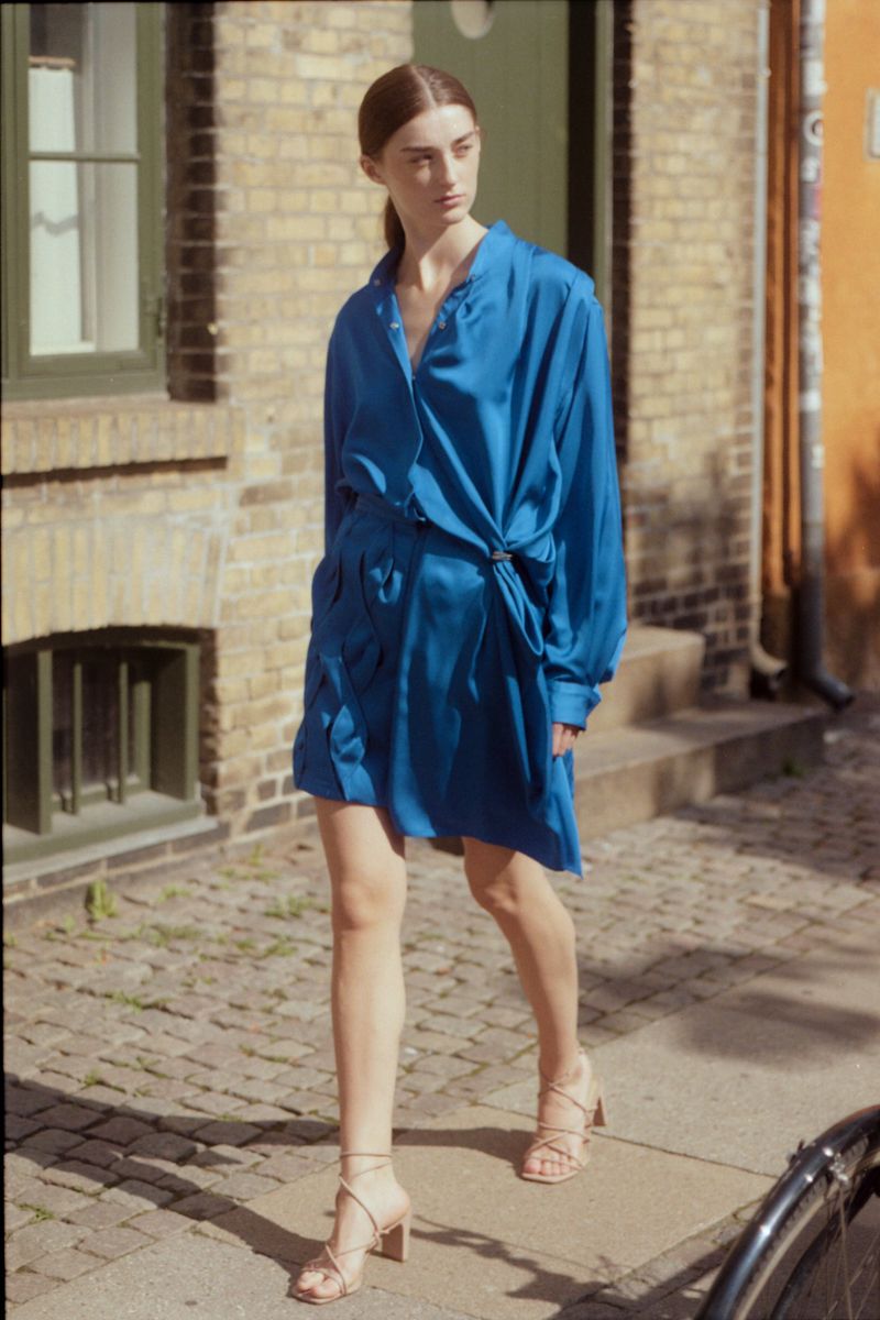 Woman in a blue dress walks on a brick sidewalk in front of a brick building wearing strappy heels, looking away from the camera.