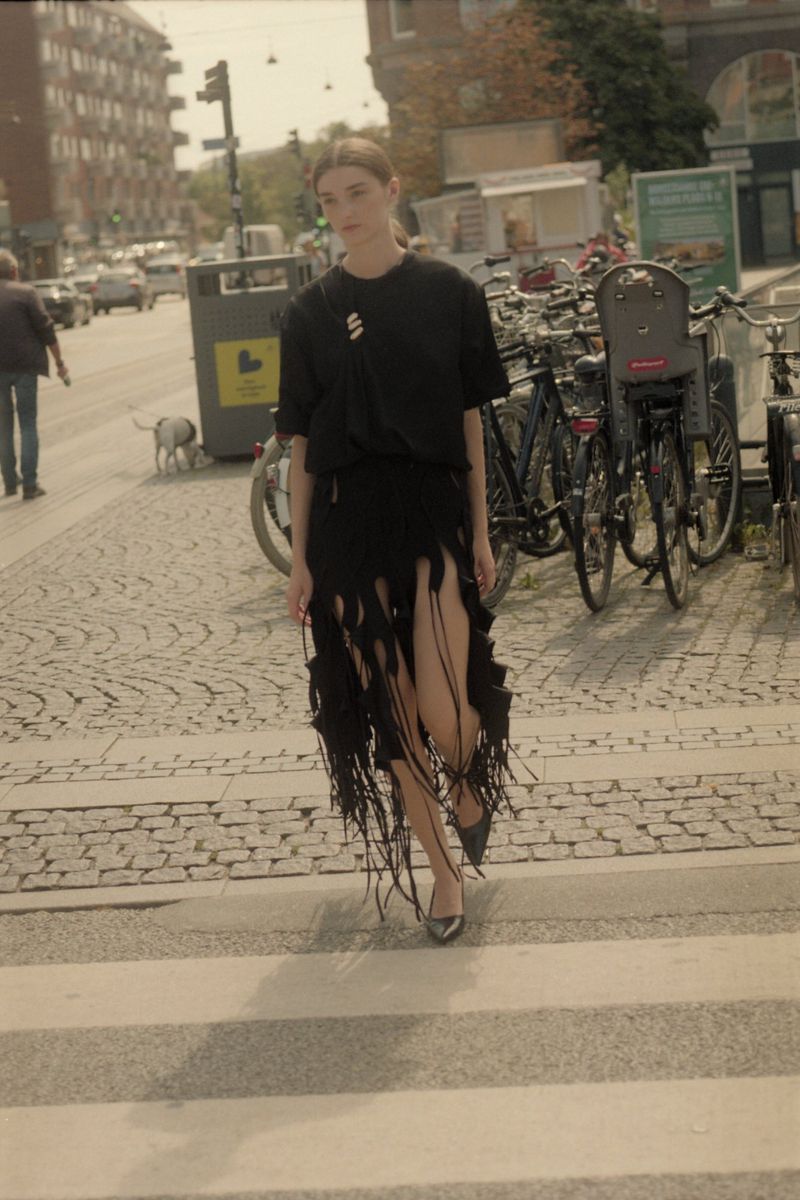 A young woman in a black dress walks across a crosswalk with bicycles parked in the background on a sunny day in an urban setting.