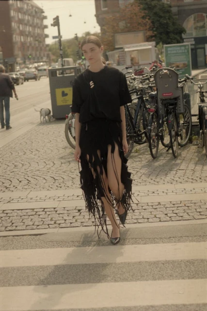 A young woman in a black dress walks across a crosswalk with bicycles parked in the background on a sunny day in an urban setting.