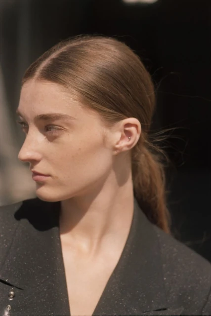 Close-up profile shot of a young woman with brown hair pulled back, wearing a sparkly black blazer, against a blurred dark background.