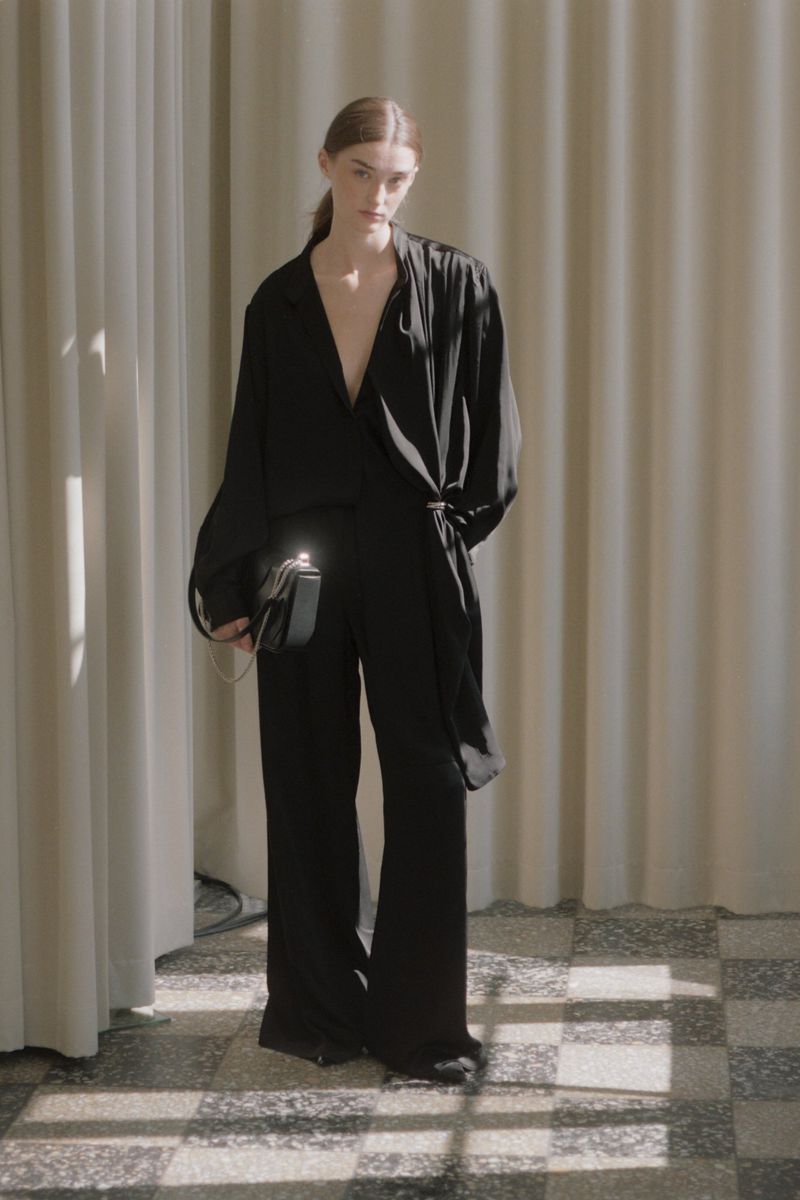 Fashionable woman in a black wrap top and trousers, carrying a rectangular black purse, posing in front of a neutral curtain.