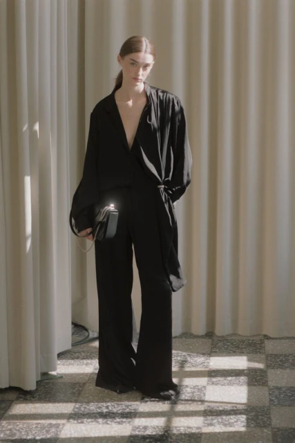 Fashionable woman in a black wrap top and trousers, carrying a rectangular black purse, posing in front of a neutral curtain.