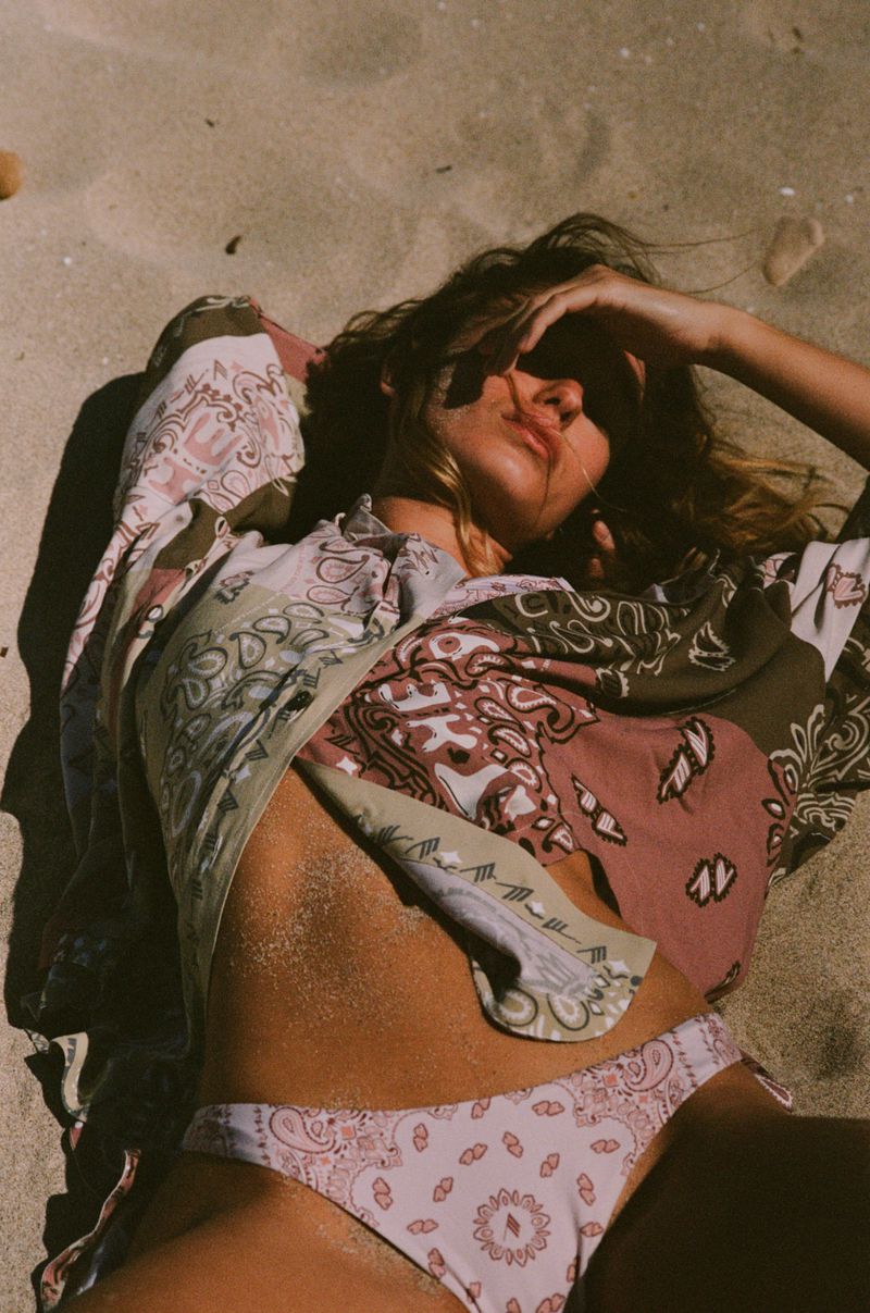 Woman in patterned bikini and shirt poses on tan sand with hand near forehead, illuminated by sunlight at the beach on a bright day.