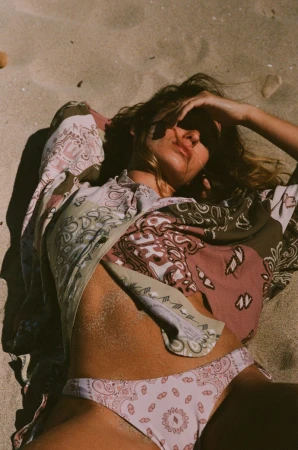 Woman in patterned bikini and shirt poses on tan sand with hand near forehead, illuminated by sunlight at the beach on a bright day.