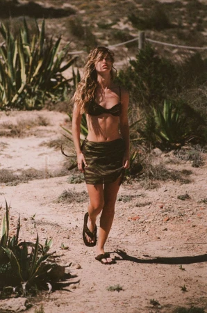 A long-haired woman in a bikini top and skirt walks on a sandy path among desert plants, captured in a warm-toned outdoor shot.