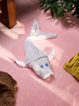 A white towel folded in the shape of a fish on a pink surface with a wooden furniture piece and green leaves in the background.