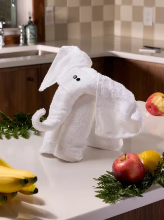 A white towel is folded into an elephant shape on a white countertop with fruit. A modern kitchen is visible in the background.