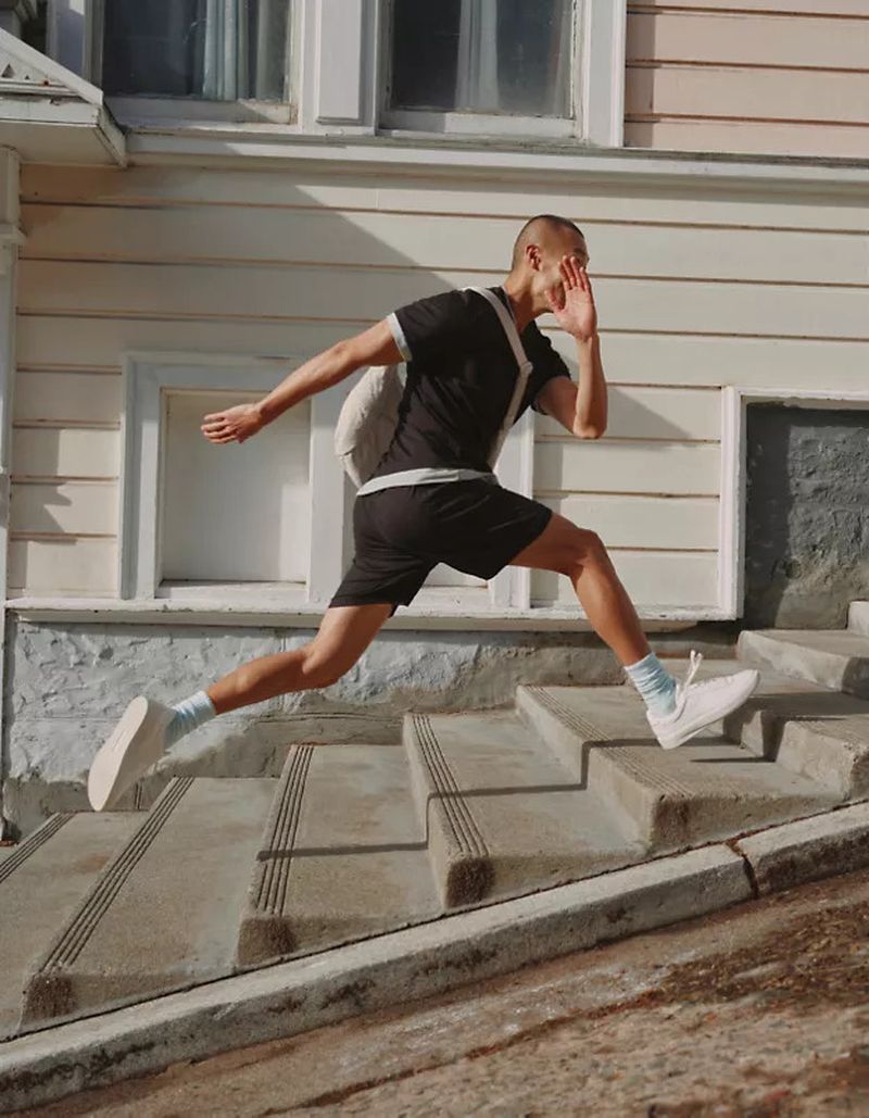 Adult male running up concrete stairs outside a white-painted building with window trim on a sunny day wearing sportswear and a backpack.