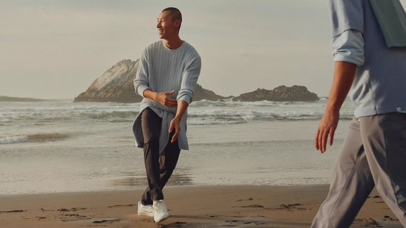 Man wearing a light blue sweater and dark pants smiling on a sandy beach near the ocean with rocky outcrops.