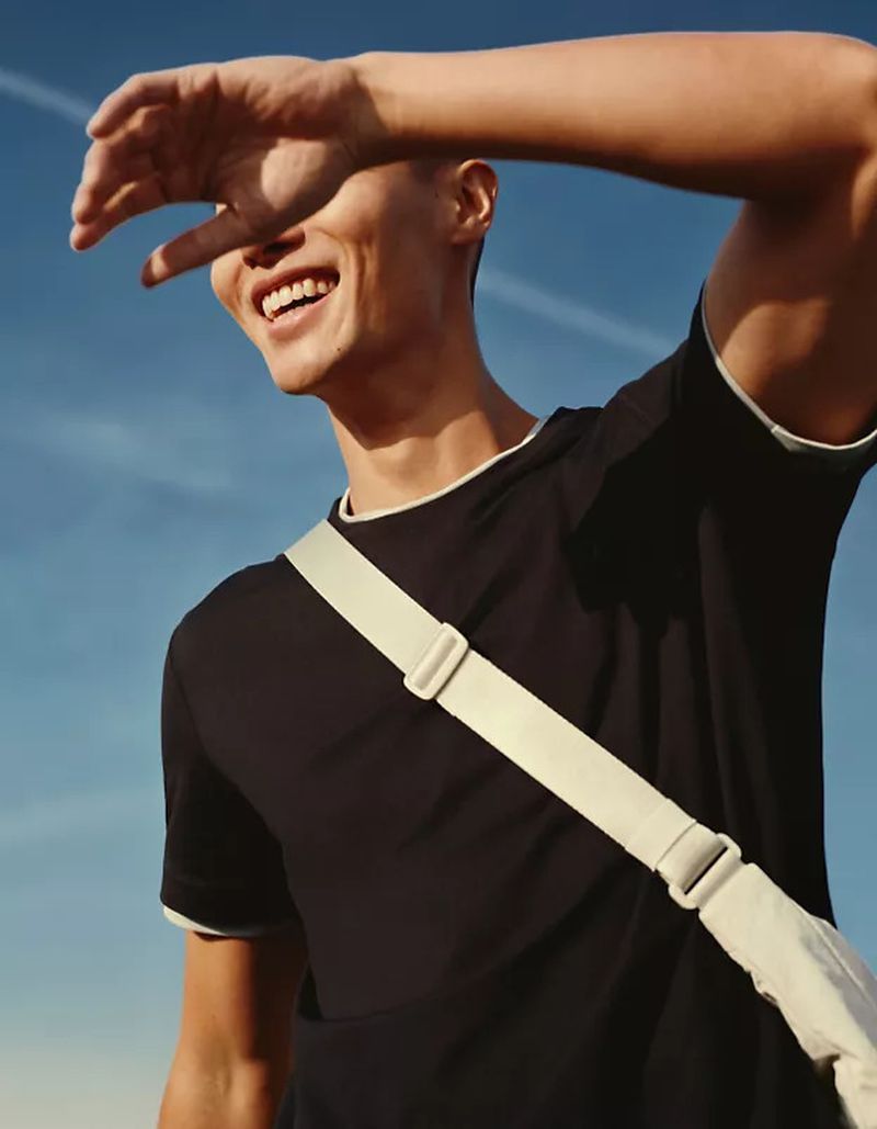 A cheerful, sunlit portrait of a smiling adult male outdoors, wearing a black t-shirt with a shoulder bag, against a blue sky.