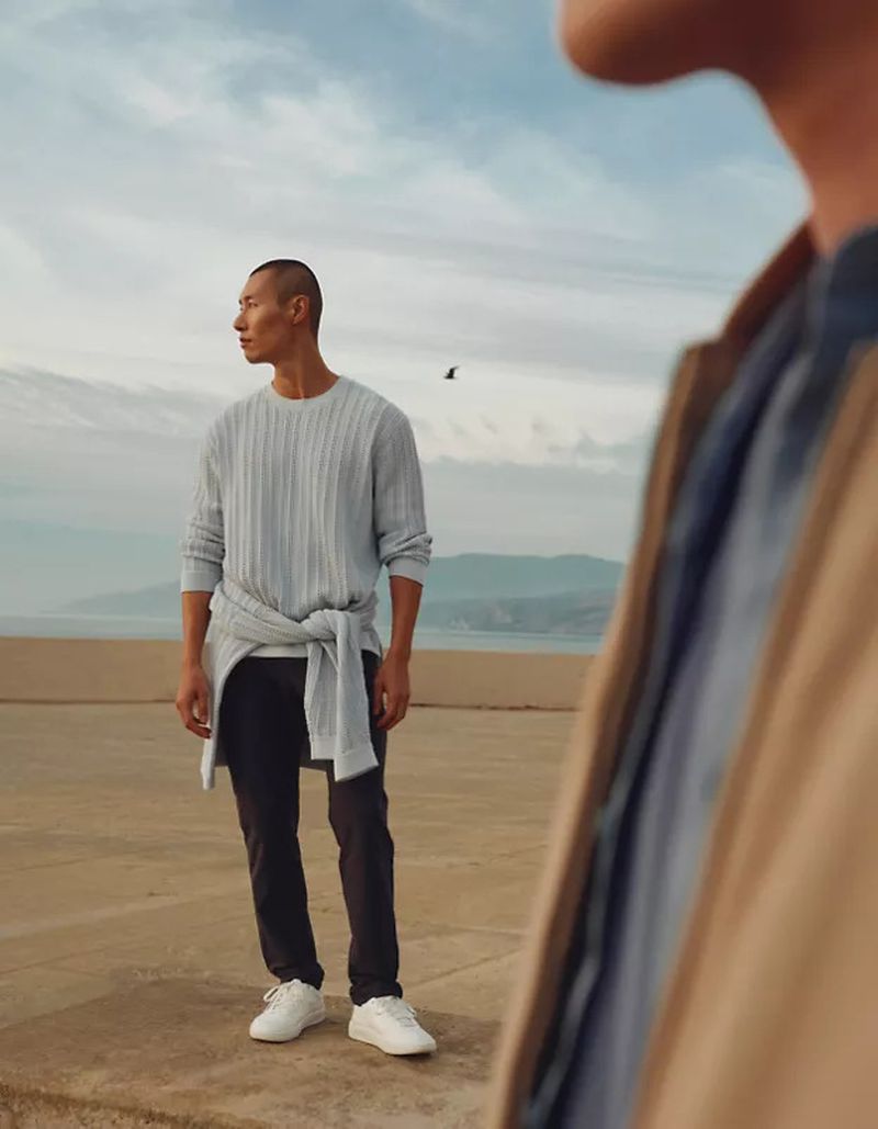 An Asian man in light-blue knitted sweater and dark pants stands on a rooftop overlooking a blurred background with water and mountains.