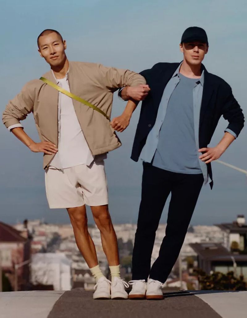 Two men in casual fashion stand arm-in-arm against blurred city backdrop. Light beige and blue color palette, daytime outdoor shot.