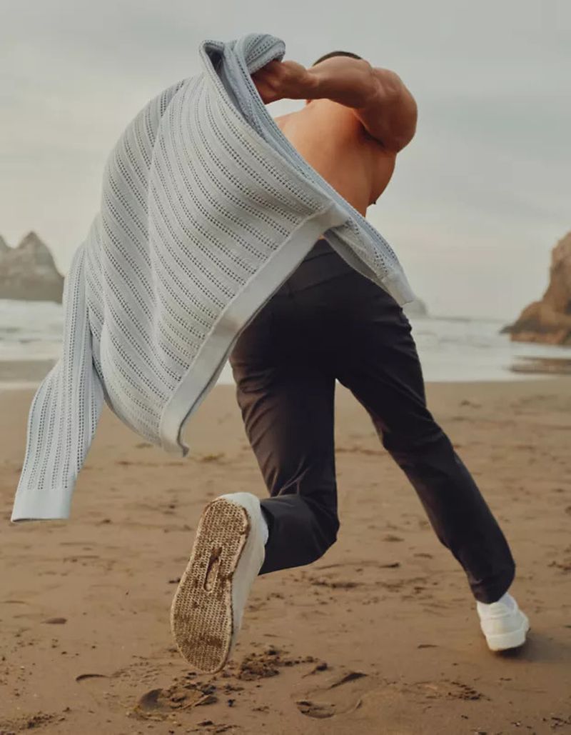 Shirtless man running on a sandy beach holding up a light grey knit sweater for Lululemon S/S 25 casual menswear campaign