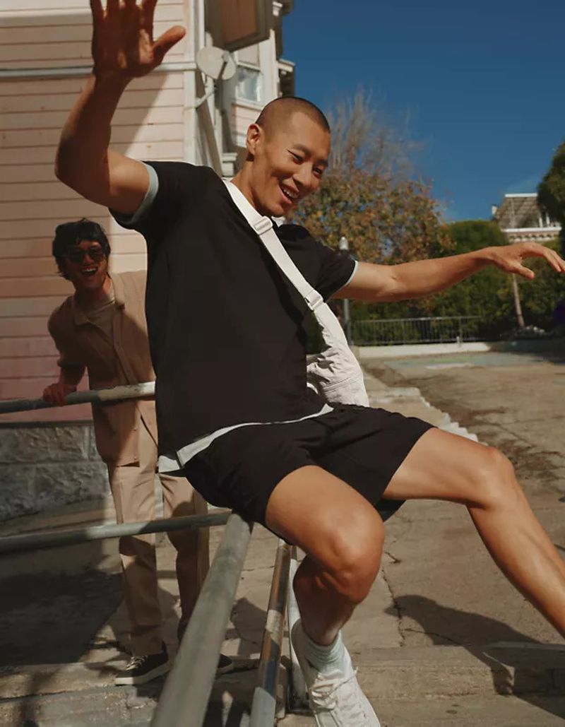 Two adult men pose on metal rails outside a pink building under a clear sky, one sitting and waving, the other leaning and smiling.