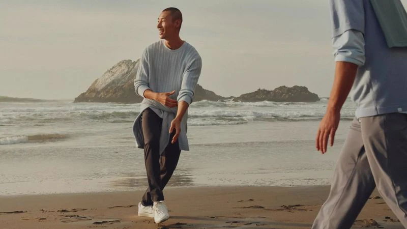 Man wearing a light blue sweater and dark pants smiling on a sandy beach near the ocean with rocky outcrops.