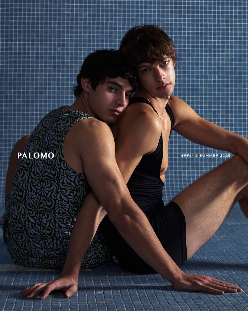 Fashion photo of two adult males sitting closely, one wearing a floral tank top, the other in a black tank top, in front of a blue tile background.