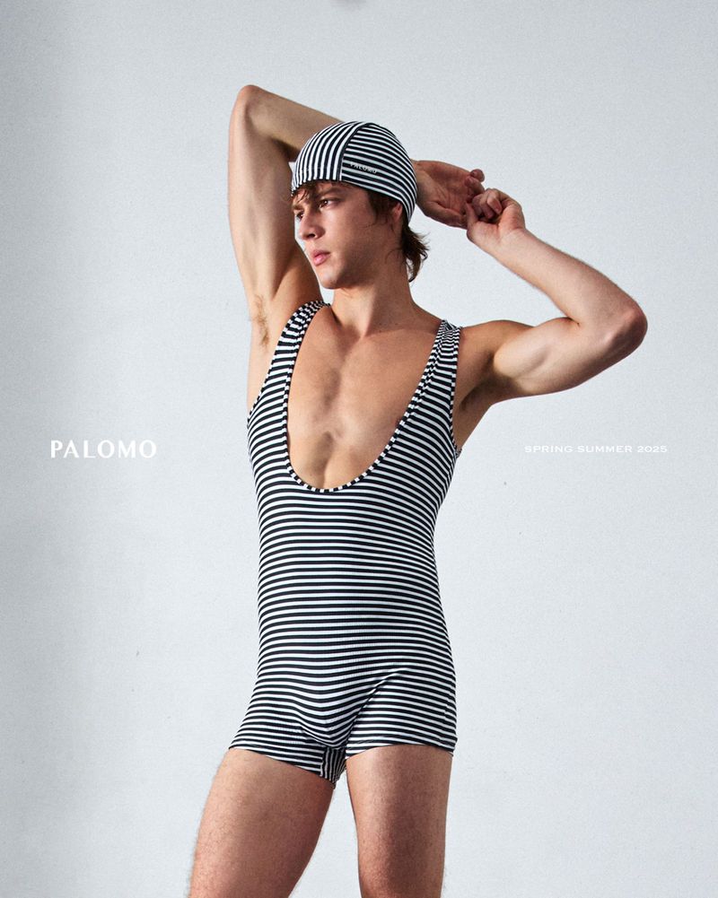 Man poses in retro striped swimsuit and cap against white backdrop. He wears a black and white striped bathing suit and swim cap.