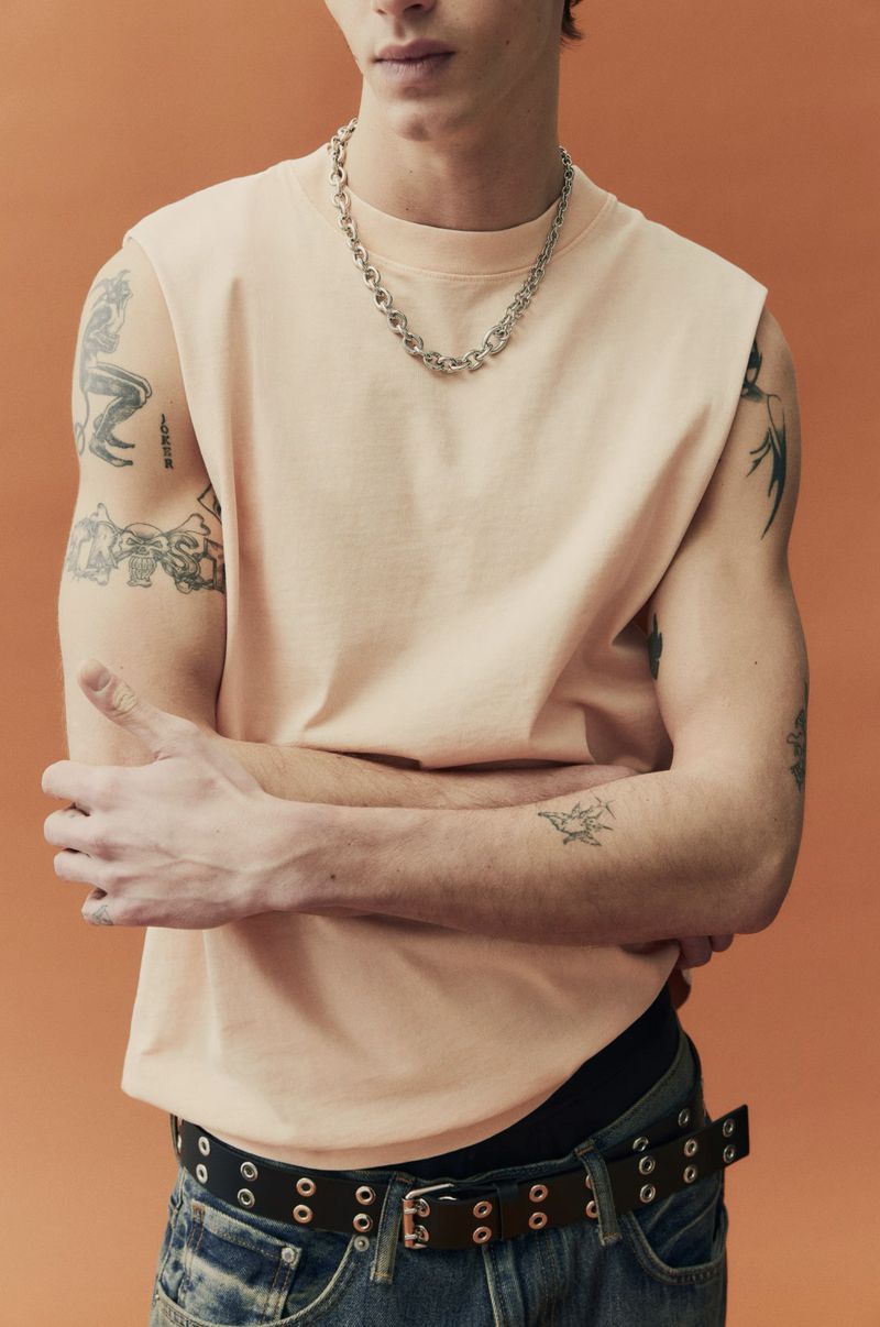 Torso of a young adult male wearing a peach t-shirt, jeans, and a silver chain necklace, posing against a peach-colored backdrop.