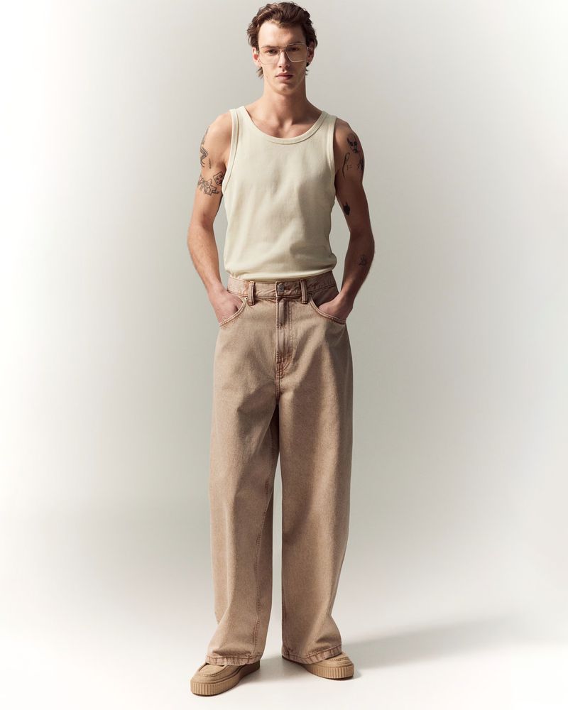A slender man with curly brown hair poses indoors, wearing a beige tank top and light brown, wide-leg jeans, standing in front of a white wall.