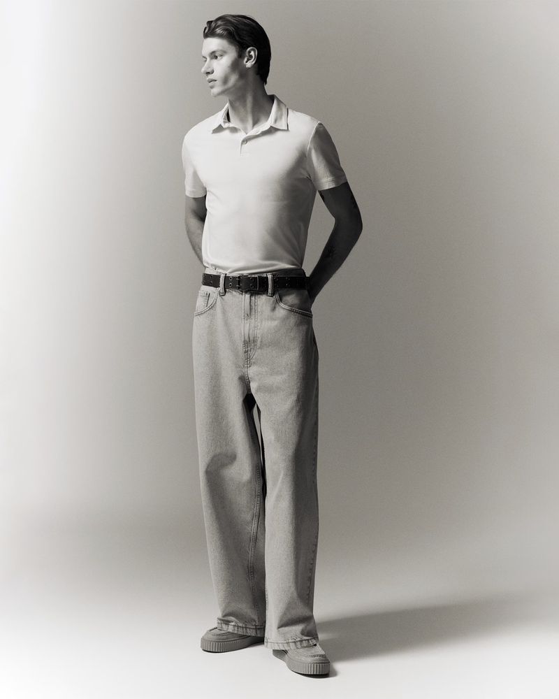 Monochrome studio shot of a young adult male model in polo shirt, jeans, and sneakers, standing with hands clasped behind his back.