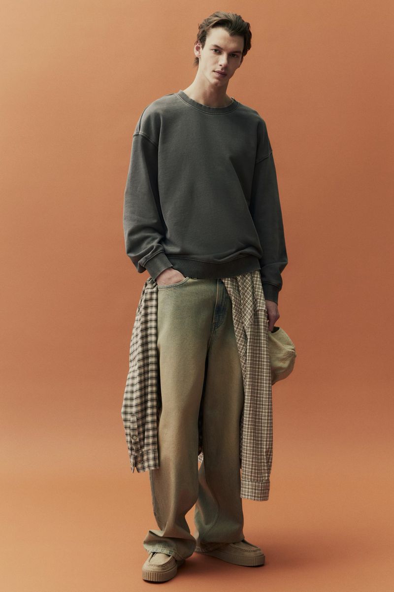 Full-length shot of a young adult male model in an olive crewneck sweatshirt, tan wide-leg jeans, and loafers, posed against an orange backdrop.