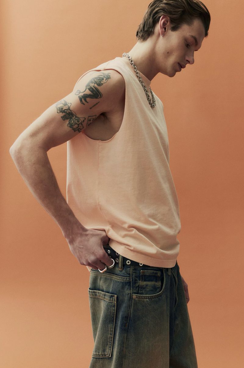 Studio shot of a young adult male model in a peach tank top and denim pants, posing against an orange background, showcasing casual fashion.