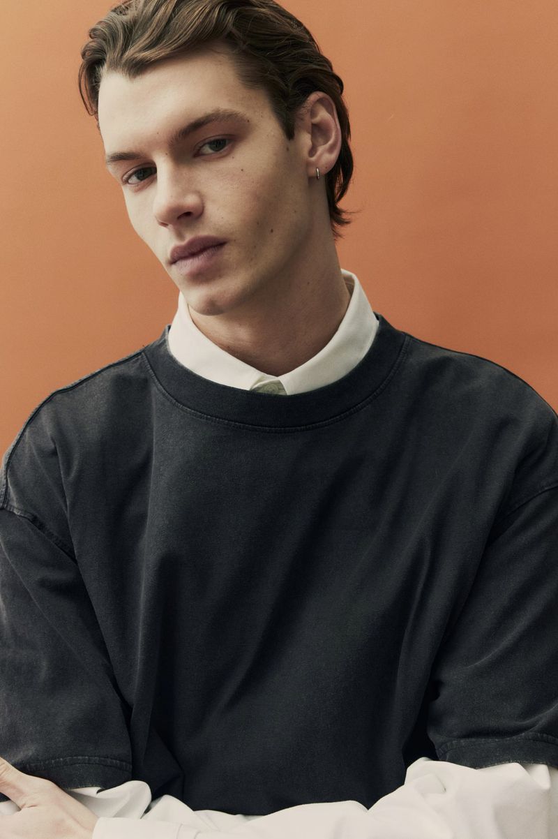Close-up portrait of a male model wearing a dark crewneck sweatshirt over a white collared shirt against an orange studio backdrop.