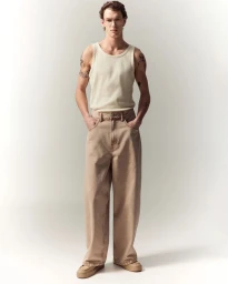 Full-length portrait of a male model wearing a ribbed cream tank top and beige trousers against a neutral background.