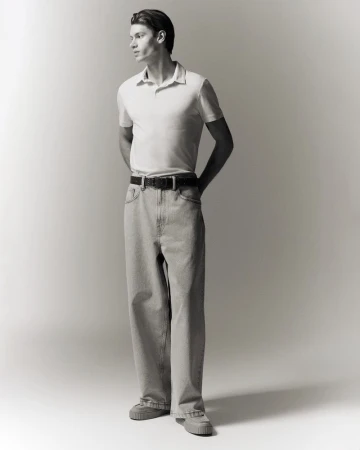 Full-length portrait of a male model wearing a white polo shirt and light grey jeans against a seamless white background.
