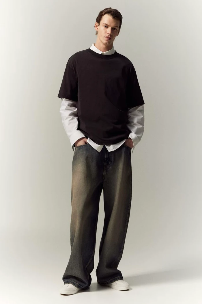 Full-length portrait of a male model wearing a layered black t-shirt over a white shirt and baggy jeans against a light background.
