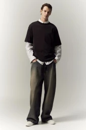 Full-length portrait of a male model wearing a layered black t-shirt over a white shirt and baggy jeans against a light background.