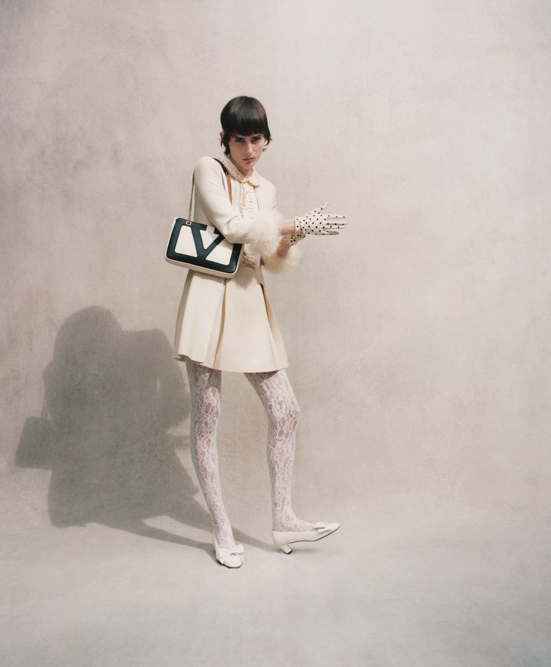 A woman in a cream-colored dress and lace tights poses in a studio with a black and white handbag and polka dot gloves.
