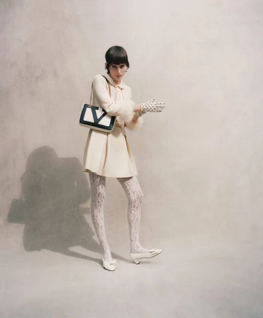 A woman in a cream-colored dress and lace tights poses in a studio with a black and white handbag and polka dot gloves.
