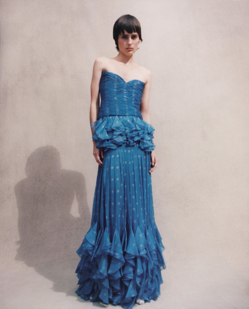 A woman poses in a strapless teal gown with ruffled detailing against a muted studio backdrop, creating a fashion-editorial aesthetic.