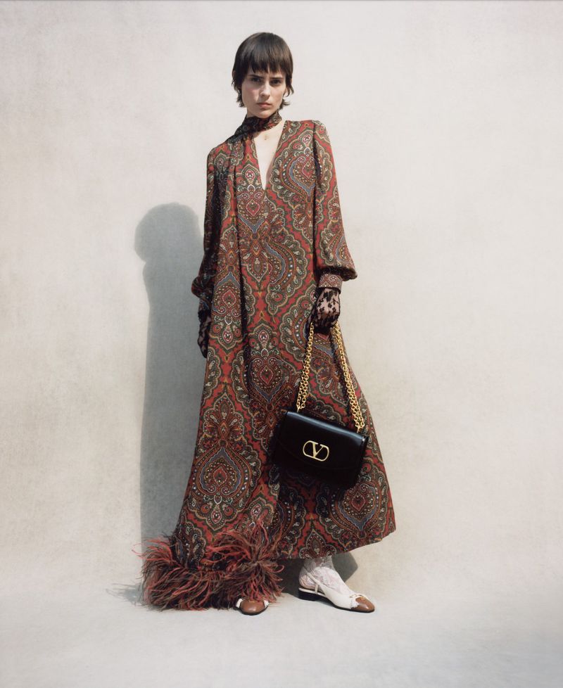 A single female model wears a patterned maxi dress and carries a black leather Valentino purse with a gold chain and V logo.