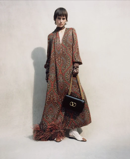 A single female model wears a patterned maxi dress and carries a black leather Valentino purse with a gold chain and V logo.