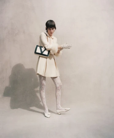 Model wearing cream dress and patterned tights holding a black and white shoulder bag against a textured background