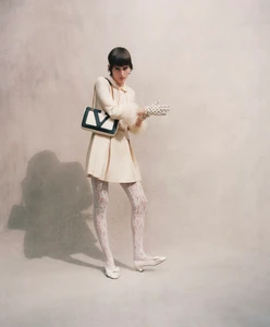 Model wearing cream dress and patterned tights holding a black and white shoulder bag against a textured background