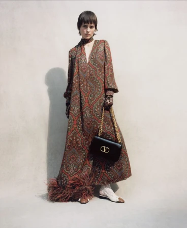 Model wearing a long paisley print dress holding a black shoulder bag against a plain wall