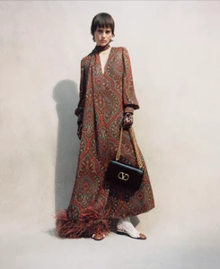 Model wearing a long paisley print dress holding a black shoulder bag against a plain wall