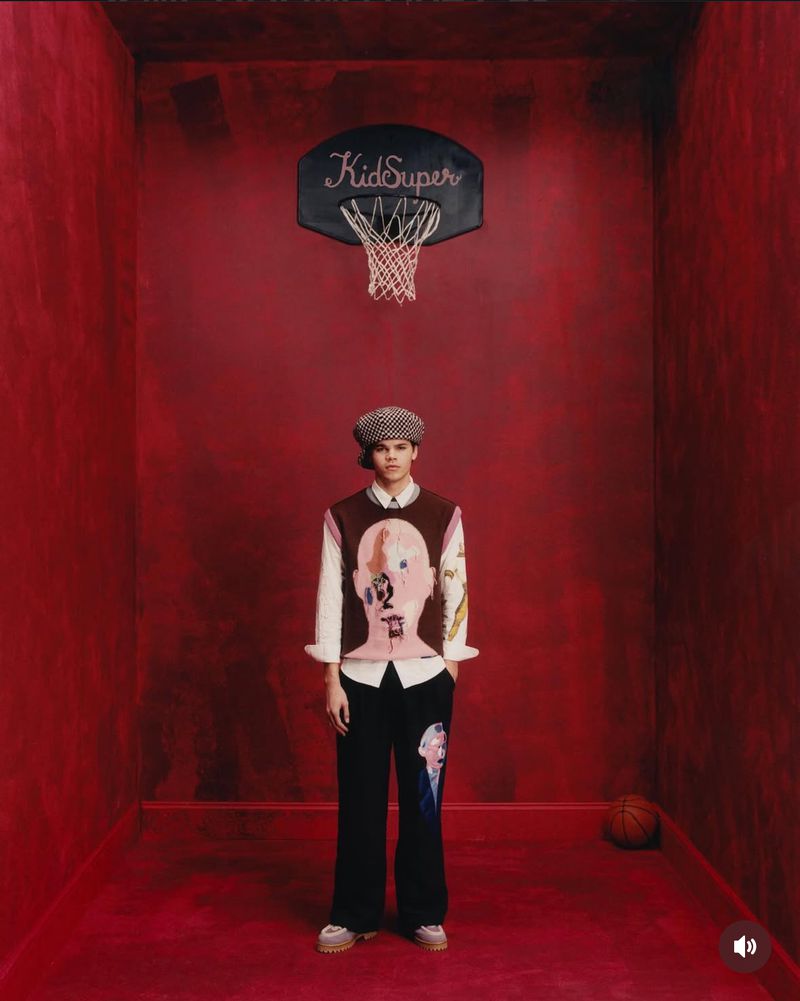 A young adult man stands in a red room wearing a printed sweater vest, black pants, and a newsboy cap with a KidSuper basketball hoop.