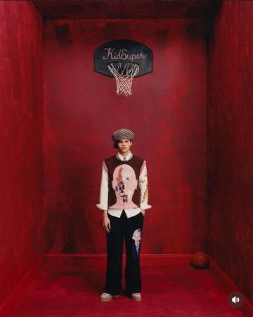 Model standing in a red room beneath a basketball hoop wearing illustrated knitwear and a cap for KidSuper