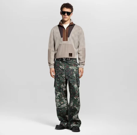 Full-shot of a model wearing a beige quarter-zip sweatshirt with brown accents, camo pants, black sunglasses, and shoes in a studio.